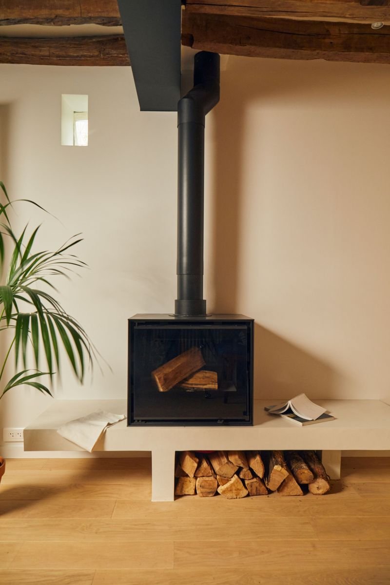 Poêle à bois noir posé sur une banquette maçonnée, sous les poutres anciennes